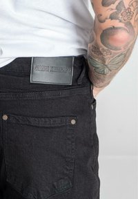 Jeans de mezclilla negra con un parche de cuero de la marca que dice "GIANNI KAVANAGH." Costuras visibles y detalles en los bolsillos traseros.