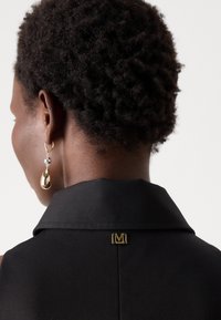 Črna brezrokavna obleka z visokim ovratnikom, ki ima zlato oznako na hrbtni strani in zlate kapljičaste uhane na modelovem ušesu.