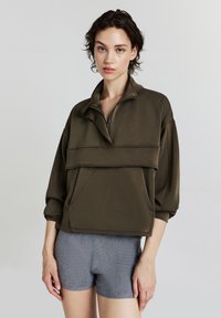 Giovane che indossa una felpa oversize verde oliva con zip e maniche a 3/4, insieme a pantaloni corti grigi in maglia, in piedi davanti a uno sfondo semplice.