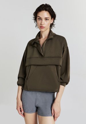 Giovane che indossa una felpa oversize verde oliva con zip e maniche a 3/4, insieme a pantaloni corti grigi in maglia, in piedi davanti a uno sfondo semplice.
