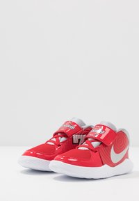 Červené atletické boty s lakovanou povrchovou úpravou, síťovaným jazykem a stříbrným logem Nike. Mají nastavitelné popruhy a texturovanou podrážku.