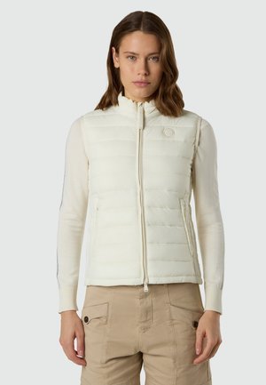 Donna che indossa un gilet trapuntato bianco senza maniche sopra una maglia a maniche lunghe e pantaloni cargo beige, in piedi davanti a uno sfondo chiaro e uniforme.