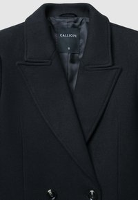 Cappotto in misto lana nero con colletto a revers, design doppiopetto e due bottoni neri in evidenza. Interno foderato in tessuto scuro.