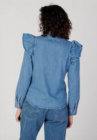Camicia in denim blu con dettagli arricciati sulle spalle, maniche lunghe con polsini a bottone e orlo curvo. Texture liscia con cuciture in evidenza.