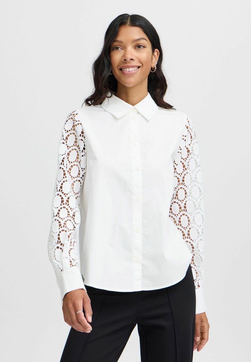 White button-up shirt with lace long sleeves featuring circular cutouts; smooth fabric, classic collar, and curved hem.