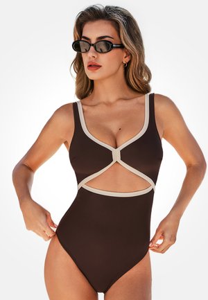Kobieta w brązowym jednoczęściowym kostiumie kąpielowym z beżowymi wykończeniami i wycięciem z przodu, poprawiająca bok, nosząca czarne owalne okulary przeciwsłoneczne.