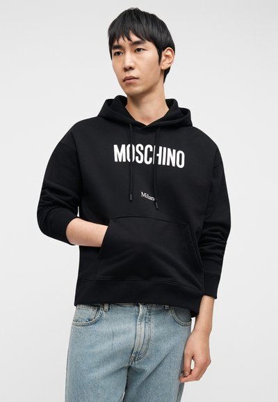 Μαύρο φούτερ με κουκούλα και το λογότυπο "MOSCHINO" σε λευκό, μπροστινή τσέπη και κορδόνια. Στυλιζαρισμένο με ανοιχτόχρωμο τζιν παντελόνι.