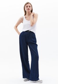 Široké tmavé džínové kalhoty s předními sklady, doplněné bílým topem bez rukávů. Modelka má na sobě černý opasek a šedé slip-on boty.