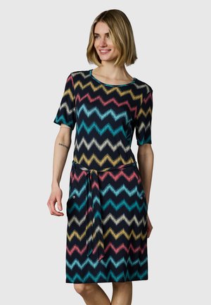 Kobieta w sukience do kolan z krótkimi rękawami i wielokolorowym wzorem w zygzak, z przewiązanym paskiem i bocznymi kieszeniami, uśmiechająca się i patrząca w prawo.