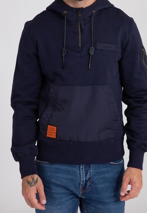 Homme portant un sweat à capuche bleu marine avec fermeture éclair frontale, cordons, écusson sur la poitrine et poche kangourou avec étiquette orange, assorti à un jean bleu.