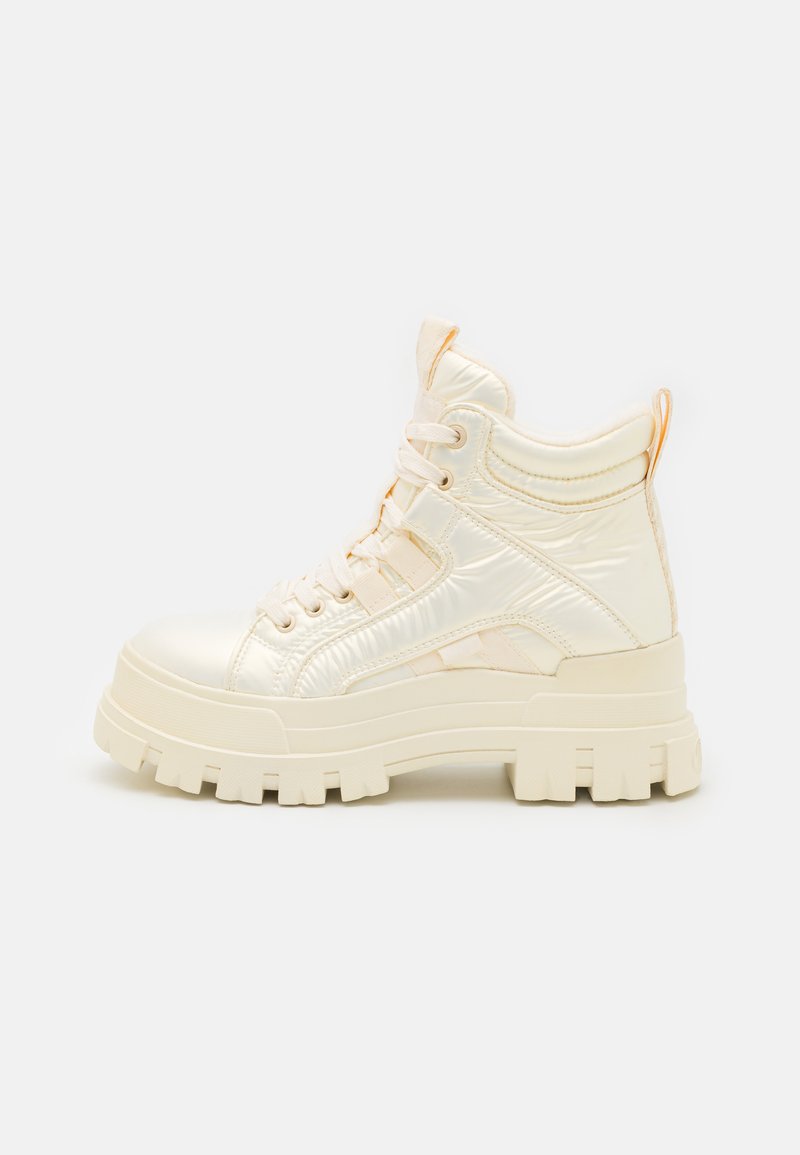Høye sneakers laget av skinnende, kremfarget syntetisk materiale med tykke, klumpete såler og teksturerte detaljer. Har snøring.