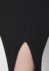 Abito nero a costine con spacco laterale, che mette in mostra un tessuto testurizzato con righe verticali. Lo spacco rivela una parte di pelle.
