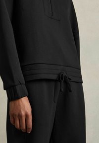 Felpa nera con tasca con zip, polsini elasticizzati e vita con cordino. Realizzata in tessuto liscio e stretch con finitura opaca.