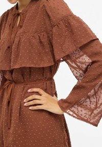 Robe marron texturée avec des manches à volants superposés, un col à nouer et une taille cintrée. Le tissu présente de petits points en relief pour plus de détails.