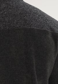 Kleidungsstück aus Wollmischung mit einem strukturierten grauen Oberteil und einem glatten schwarzen Unterteil. Weicher, warmer Stoff mit leichtem Glanz.