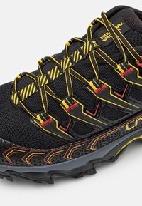 Sapatilha desportiva preta com parte superior em malha, detalhes vibrantes em amarelo e vermelho, e uma sola texturizada com padrões geométricos para aderência.