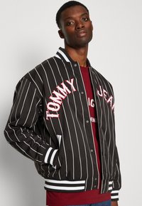 Mustriga musta bomber-jakk, millel on valge ja punane tikitud "TOMMY JEANS" tekst, soonikkoes käised ja krae ning küljetaskud.