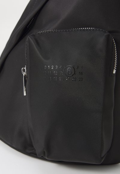Svart tygryggsäck med en dragkedjeficka som har präglade siffror och en logotyp. Slät textur och minimalistisk design med silverfärgad hardware.