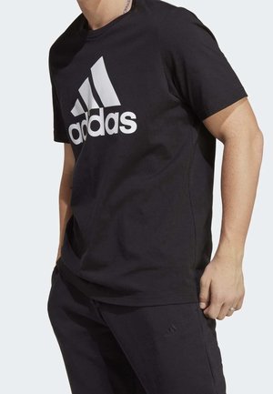 Osoba nosząca czarną koszulkę Adidas z dużym białym logo oraz czarne spodnie z małym logo Adidas.