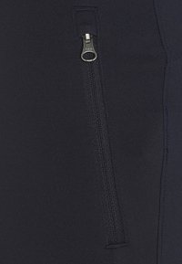 Los pantalones deportivos de color azul marino presentan un material elástico texturizado y un bolsillo con cremallera en el costado, añadiendo un detalle funcional al diseño.
