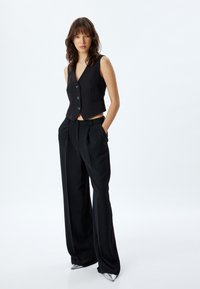 Gilet nero senza maniche con scollatura a V e cinque bottoni, abbinato a pantaloni neri a vita alta e gamba larga e scarpe metalliche a punta.