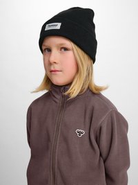 Ung person med skulderlangt blondt hår iført en sort beanie og en brun fleecejakke med lynlås med et bi-logo på brystet.