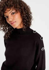 Pull en tricot noir avec un col haut en côtes, orné de boutons argentés et noirs sur les épaules. Texture douce, design ajusté.