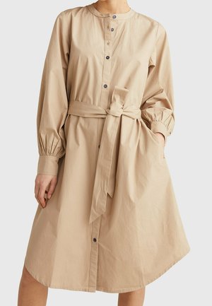 Beżowa sukienka na długi rękaw z zapięciem na guziki z przodu, marszczonymi mankietami, okrągłym kołnierzem, wiązaną kokardą w talii i boczną kieszenią, noszona przez stojącą osobę.