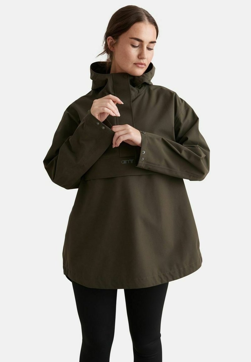 Femme ajustant le poignet d'une veste de pluie à capuche vert foncé, portant un pantalon noir, sur un fond clair uni.