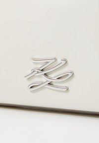 Kremfarget teksturert lærprodukt med en skinnende sølvfarget metalllogo med elegante kurver og spiraler. Enkle design detaljer er til stede.