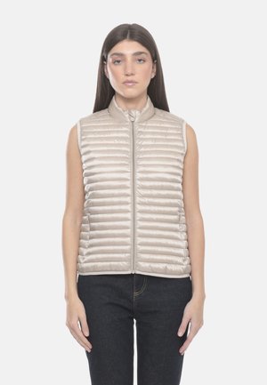 Donna che indossa un gilet trapuntato beige senza maniche con zip frontale, abbinato a pantaloni neri, in piedi su uno sfondo semplice.