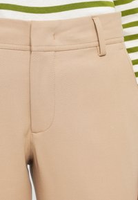 Gros plan sur la taille d’un pantalon beige avec passants de ceinture porté sur une chemise blanche à rayures vertes.