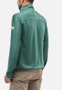 Grüne, strukturierte Fleecejacke mit hohem Kragen und Logo auf dem linken Ärmel, kombiniert mit hellen Hosen, die eine lässige Passform zeigen.