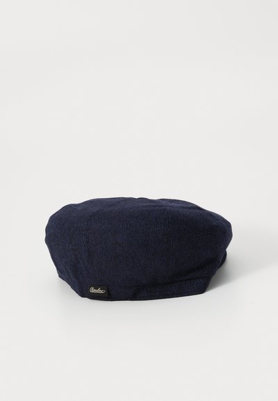 Pălărie plată din tweed în carouri herringbone de culoare bleumarin, cu o mică etichetă de brand neagră și aurie pe lateral, expusă pe un fundal alb.