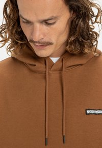 Brown hoodie in soft fabric, with a kangaroo pocket, drawstrings, and a small black logo patch on the chest.