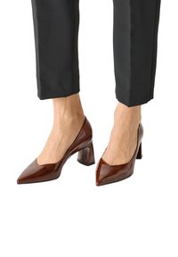 Chaussures à talons carrés en cuir verni marron à bout pointu portées avec un pantalon noir à la coupe courte, pieds visibles depuis le mollet sur un fond blanc.