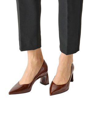 Brown patent leather pointed-toe block heels worn with black cropped pants, feet shown from mid-calf down on white background.