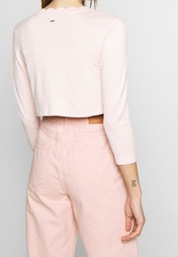 Avskuren, ljusrosa stickad tröja med långa ärmar och vågig halsringning, tillsammans med högmidjade, ljusrosa byxor med en läderetikett.