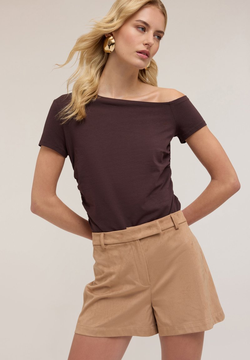 Tricou maro cu umerii goi, asociat cu pantaloni scurți kaki, având o textură netedă și o croială ajustată, cu talii și buzunare laterale.