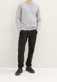 Uomo che indossa un maglione a maniche lunghe grigio chiaro a trama, pantaloni neri cuffed con grandi tasche e stivali neri con lacci, in piedi con le mani nelle tasche.