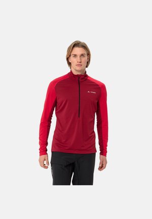 Mann trägt ein rotes langärmeliges Sportoberteil mit schwarzem Reißverschluss und schwarze Hose, steht vor einem schlichten hellen Hintergrund.