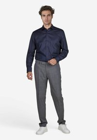 Marineblaues Button-up-Hemd aus glattem Stoff, kombiniert mit grauen karobierten Hosen und weißen Sneakers. Das Model steht aufrecht, die Hände lässig platziert.
