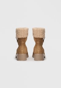 Botas de tobillo de ante marrón con un puño de pelo sintético tipo shearling en color crema y textura, con un tacón bajo y un detalle de correa lateral.