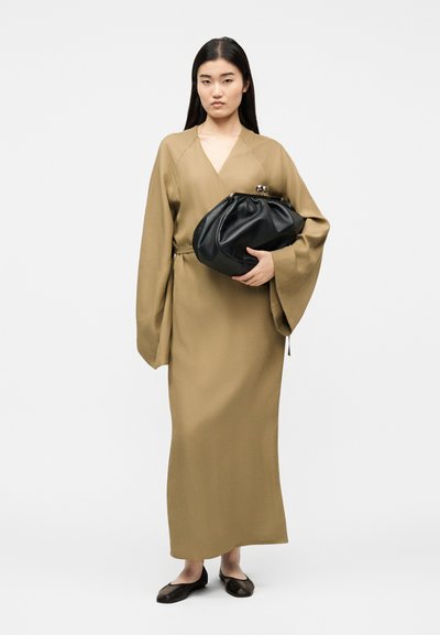 Femme debout portant une longue robe portefeuille beige clair à manches larges, tenant une grande pochette noire et portant des chaussures plates noires.
