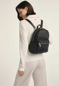 Czarny, miękki plecak z zaokrąglonym kształtem. Oferuje złote zamki, przednią kieszeń oraz regulowane paski. Nosi się na plecach.