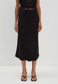 Schwarzer Midirock mit glatter Textur, der leicht ausgestellt ist und seitliche Schlitze aufweist. Kombiniert mit schwarzen Sandalen mit Riemchen.