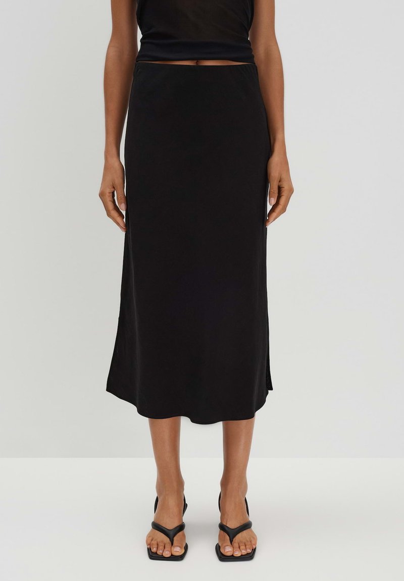 Schwarzer Midirock mit glatter Textur, der leicht ausgestellt ist und seitliche Schlitze aufweist. Kombiniert mit schwarzen Sandalen mit Riemchen.