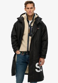 Abrigo negro impermeable con capucha, superpuesto a un suéter de polar crema y una camisa de cuadros. Presenta cremallera y un gran logo blanco en el lado.