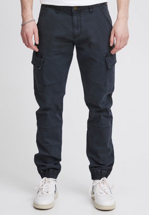 Mann in dunkelgrauen Cargo-Jogginghosen mit elastischen Bündchen und weißen Sneakers, stehend vor einem schlichten Hintergrund.