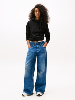 Black hooded sweatshirt, cropped fit; blue wide-leg jeans, faded, with a cut-out detail; worn with white and black sneakers.